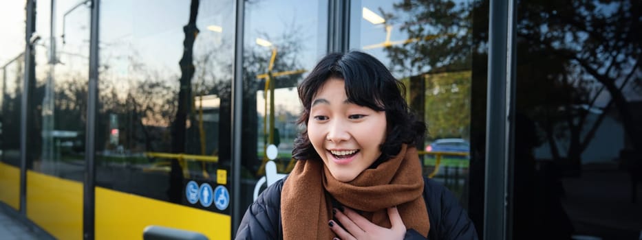 Portrait of cheerful asian girl talks on mobile phone, video chats, looks amazed at smartphone camera, stands on bus stop.
