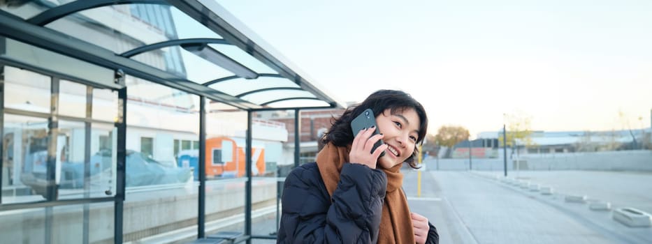 Beautiful smiling korean girl, waiting on bus stop, using public transport, talking on mobile phone, going somewhere in city.