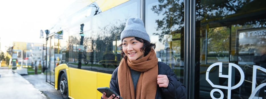 Portrait of girl standing near bus on a stop, waiting for her public transport, schecks schedule on smartphone application, holds mobile phone, wears warm clothes.