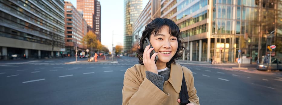 Smiling asian girl makes a phone call, stands on an empty street, calling someone on telephone, waiting for friend in city, going to a meeting.