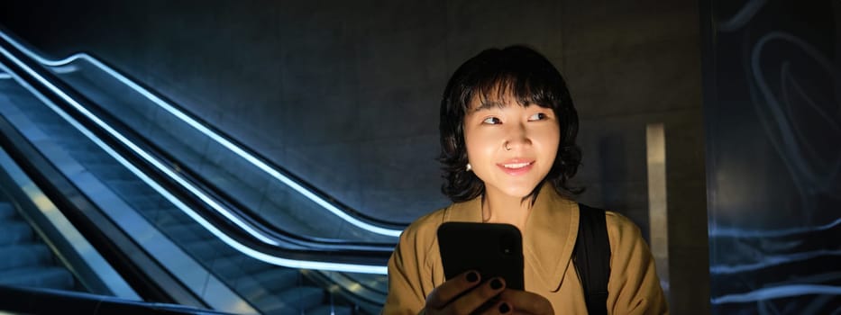 Stylish Japanese woman in headphones, standing with smartphone in hands near escalator, travelling and commuting in city, urban lifestyle concept.