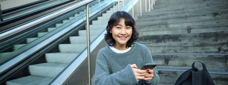 Stylish young girl student, sits on stairs with smartphone and backpack, laughs and smiles, texts message, uses social media.