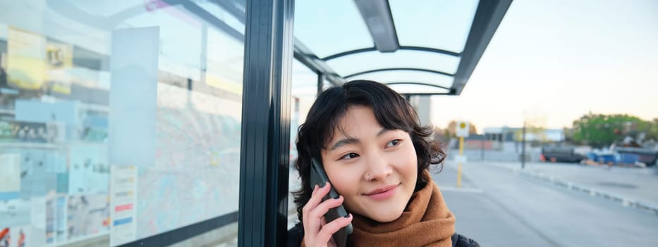 Close up portrait of cute young woman calling someone, waiting on bus stop with smartphone, using telephone while expecting public transport to arrive.