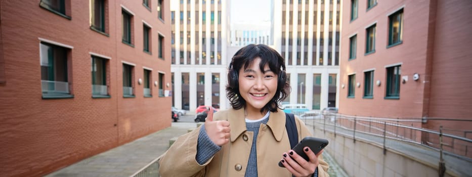 Portrait of cheerful asian girl in headphones, stands on street and shows thumbs up, found something online on her smartphone, using mobile phone.