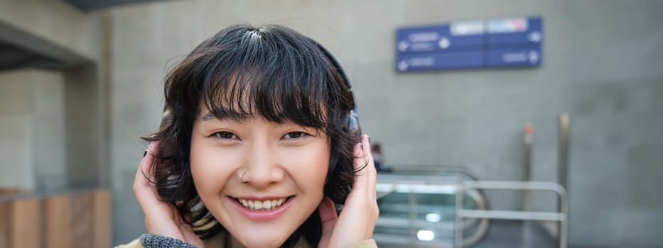 Close up portrait of stylish korean girl standing on street, listening music in headphones and smiling, posing in city centre.