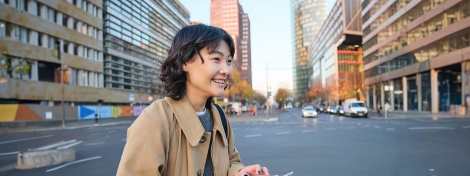 Portrait of young korean student, girl walks in city with digital tablet, stands on street, uses her gadget outdoors.