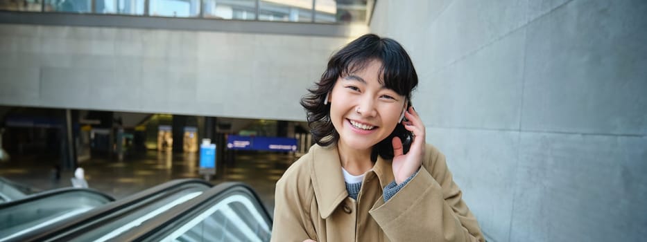 Portrait of cute smiling korean girl, goes up escalator, listens music in wireless earphones and uses mobile phone, holds smartphone.