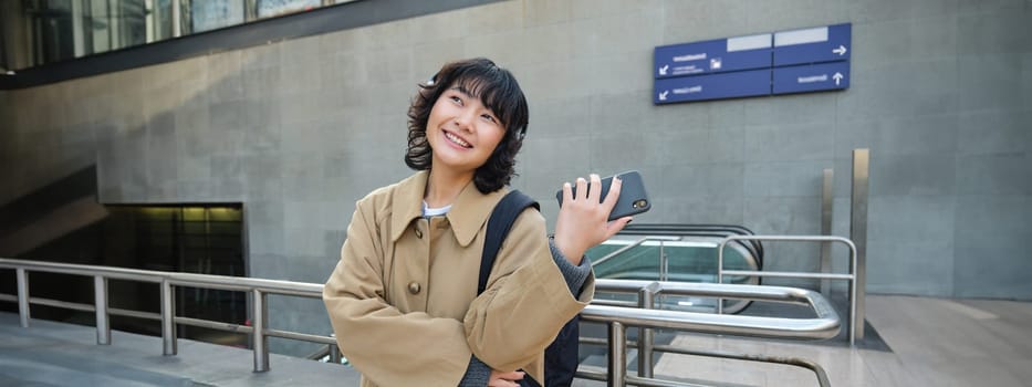 Happy young woman in headphones, stands on street with smartphone and drinks coffee, waits for commute train or bus, travels in city.