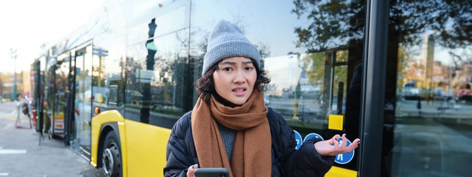 Portrait of confused young woman, looking at her smartphone app with shocked and disappointed face, shrugs shoulders, reads bad news, stands on bus stop near public transport.