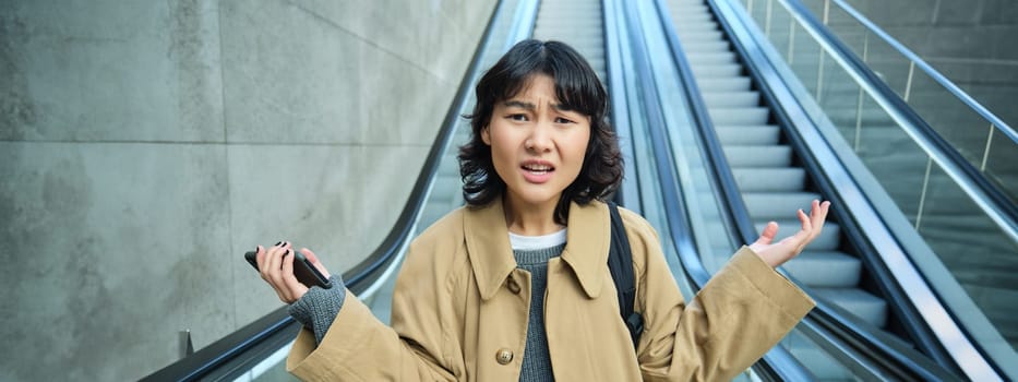 Portrait of confused asian girl doesn't know where she is, lost in unfamiliar city, goes down escalator with frustrated face, shrugging and looking at camera.
