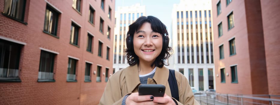 Portrait of cute girl tourist, korean woman in headphones, looks at mobile phone, uses smartphone app, map or text messages, listens music in headphones.