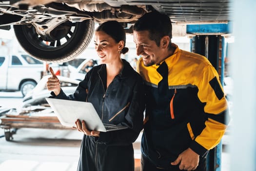 Two vehicle mechanic working together underneath lifted car, conduct car inspection with laptop. Automotive service technician in uniform carefully make diagnostic troubleshooting. Oxus