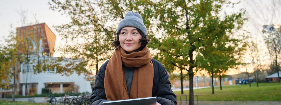 Smiling young girl, korean student sits in park in scarf and hat on chilly day, draws outdoors on digital tablet with graphic pen, makes scatches.