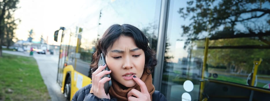 Perplexed asian girl, talks on mobile phone with concerned, thoughtful face, stands near bus stop and frowns, thinking while listening telephone conversation.