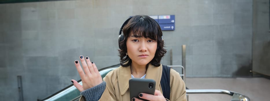 Portrait of asian girl in headphones, looks complicated at smartphone screen, puzzled by text message or notification, stands on street and shrugs.