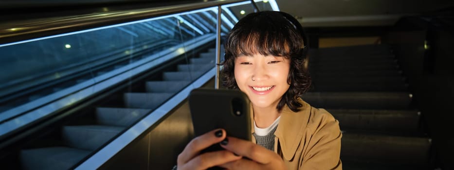 Portrait of happy asian girl in headphones, smiling and looking at smartphone, watching video or listening music on mobile phone, sitting on staircase.