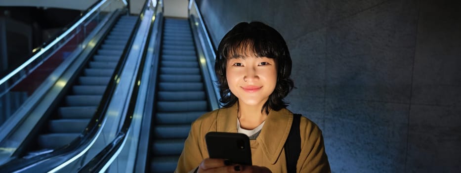 Young Japanese girl standing with smartphone, going down escalator with mobile phone, commuting in city, using public transport.