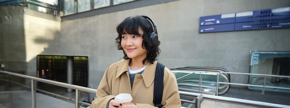 Portrait of young student, girl in headphones, drinks coffee, stands on street with backpack, commutes to university or college, smiles happily.