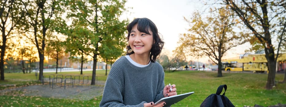 Young korean woman sits in park, draws on digital tablet, writes in gadget with pen tool, looks around and makes notes, creates artwork or scatches.