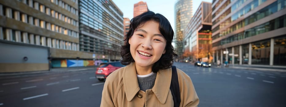 Portrait of asian girl student, stands in city centre with cars on busy street, holds digital tablet and smiles at camera.