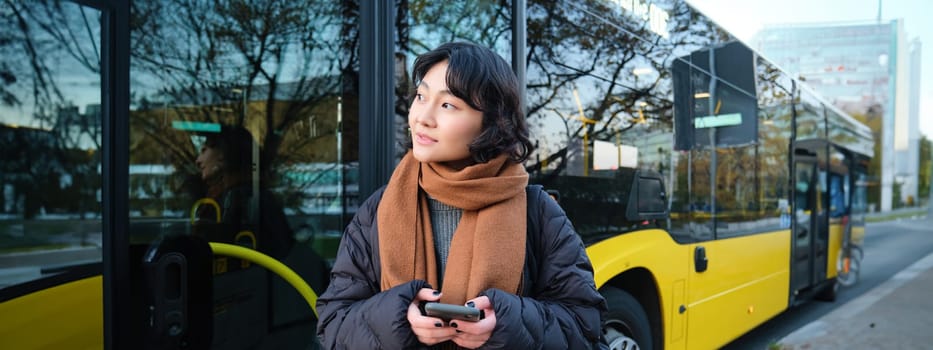 Portrait of korean girl buying ticket for public transport online, using mobile application on bus stop, wearing winter clothes.