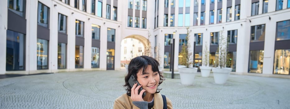 Cellular technology. Young korean woman talks on mobile phone, makes a phone call on her way home, walks down street, city centre, has telephone conversation.