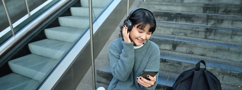 Portrait of smiling korean girl in headphones, uses smartphone and sits on stairs in mall, watches video on mobile phone. Copy space