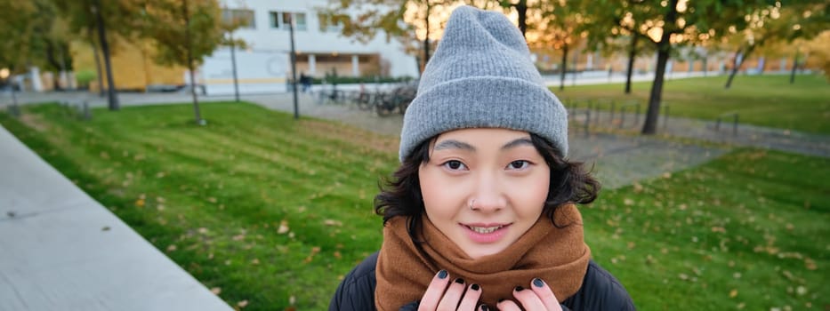 Portrait of cute asian girl in hat and scarf, walks around town in chilly spring weather, smiles and looks happy, sits in park near green grass.