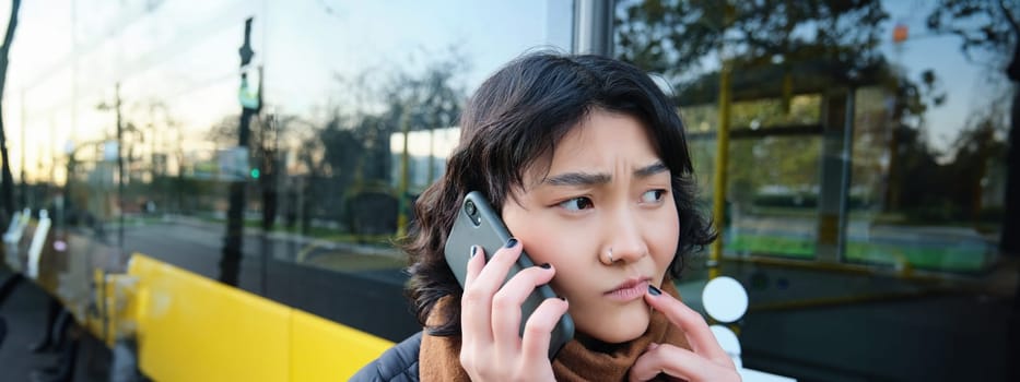 Perplexed asian girl, talks on mobile phone with concerned, thoughtful face, stands near bus stop and frowns, thinking while listening telephone conversation.