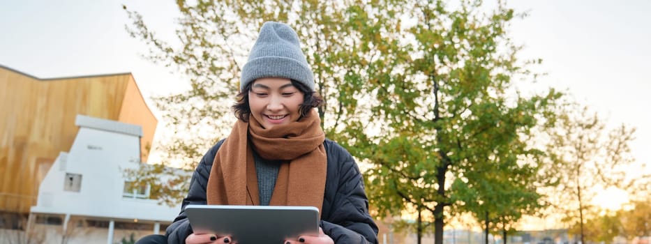 Portrait of asian girl in warm clothes, sits on bench with digital tablet and graphic pen, smiling happily, draws outdoors in chilly weather.