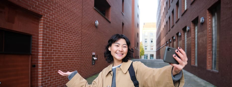 Happy korean girl, tourist takes photos on street, shows smth while records video of herself on smartphone, posing near building and smiling.