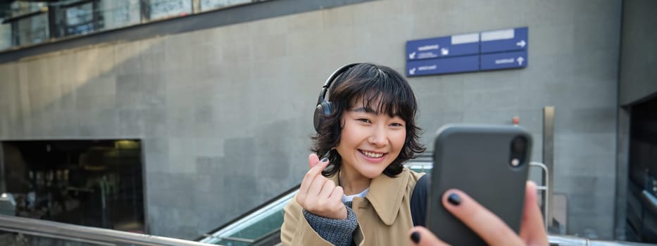 Cute and stylish Korean girl, wears headphones, takes selfie on smartphone, tourist records video or makes a photo, stands on street.