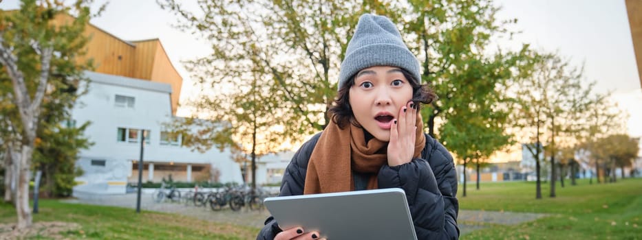 Portrait of asian girl with shocked, surprised face, holds tablet, read or watched big news, looks amazed, sits in park on bench, wears warm clothes.