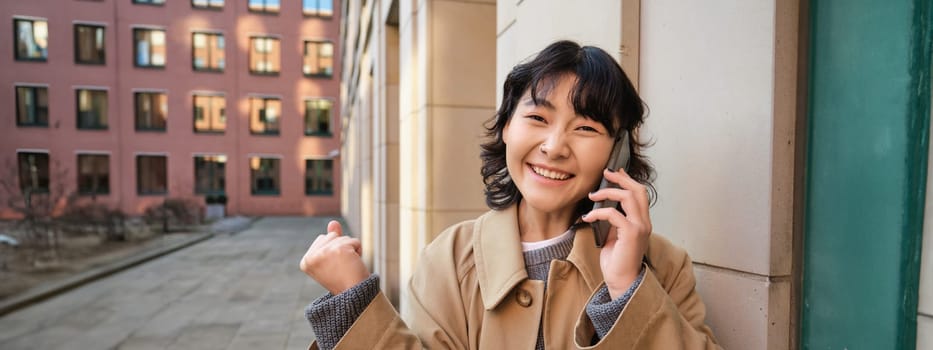 Portrait of smiling happy korean girl, stands on street, receives good news over phone, talks on smartphone and makes fist pump, celebrates, feels excitement.