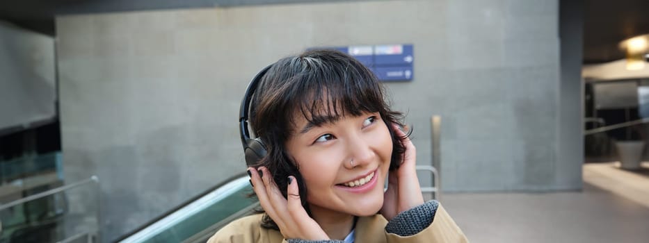 Beautiful smiling korean girl in headphones, wondering around town, standing on street and smiling, listening to music.