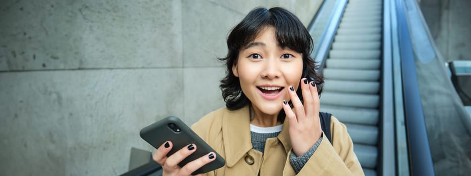 Joyful and positive korean girl, celebrates, looks surprised, goes down escalator with smartphone and looks amazed by smth.