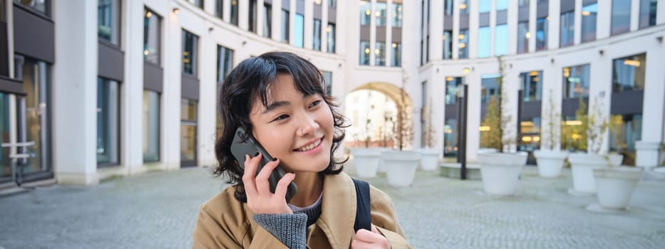 Cellular technology. Young korean woman talks on mobile phone, makes a phone call on her way home, walks down street, city centre, has telephone conversation.