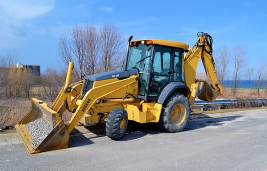 A yellow backhoe sitting on the street in Stoney Creek, Ontario close to the lake Ontario, on a beautiful sunny day in early spring.