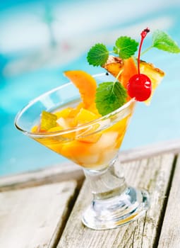 Refreshing colourful tropical fruit cocktail appetiser in a conical glass filled with pieces of fresh mixed fruit on decking alongside a blue pool