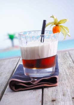 Layered alcoholic cocktail with gooseberry garnish served on a wooden deck by the sea