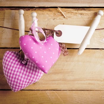 Two romantic pink patterned textile hearts hanging from rustic wooden pegs with a blank label