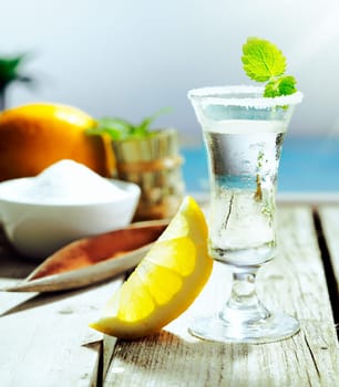 Chilled glass of vodka cocktail garnished with frosting and mint and served with lemon alongside a turquoise pool