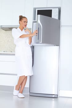 Vivacious woman in a white bathrobe and slippers standing laughing as she opens the freezer unit of her refrigerator