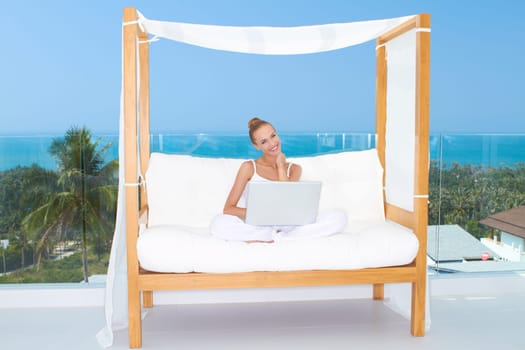 Fresh bright portrait of a woman sitting cross legged on a canopied seat with her laptop backed by a large window overlooking the ocean