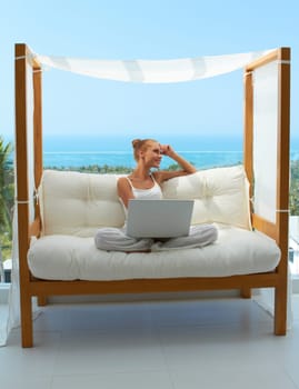 Beautiful woman sitting on a canopied seat on her patio resting her head on her hand as she takes a break from working on her laptop