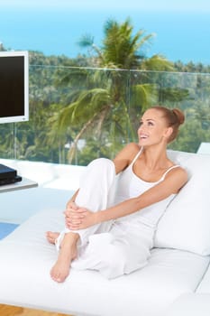 Beautiful happy woman sitting barefoot on a white sofa in her living-room with a tropical view through the large glass window