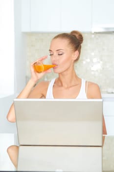 Healthy happy woman drinking from glass of fresh orange juice seated at a table using her laptop