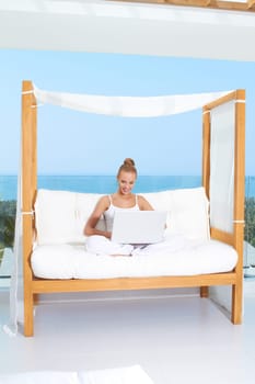 Fresh bright portrait of a woman sitting cross legged on a canopied seat with her laptop backed by a large window overlooking the ocean