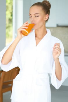 Woman in a white bath robe with her eyes closed enjoying a large glass of fresh orange juice in the morning for breakfast