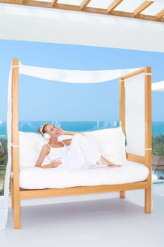 Woman reclining on a white canopied seat in the summer sunshine on the patio enjoying music on her headphones with an ocean backdrop
